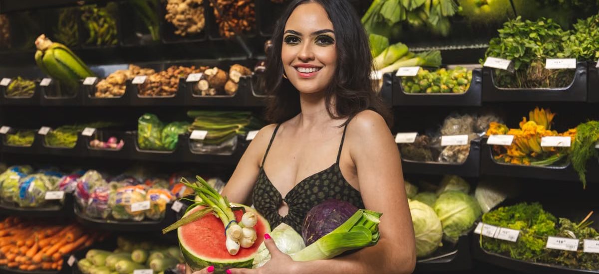 Smiling woman holding vegetables and fruit in a grocery store produce aisle.